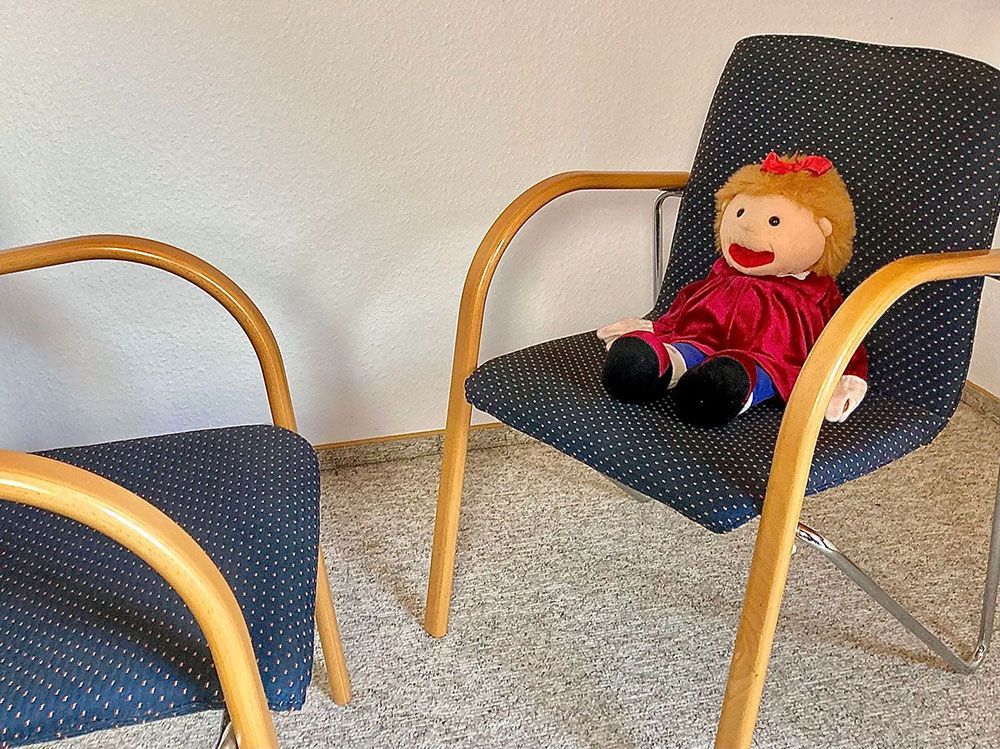 Eine Puppe sitzt auf einem blau gepolsterten Stuhl neben einem leeren Stuhl in einem Wartezimmer.