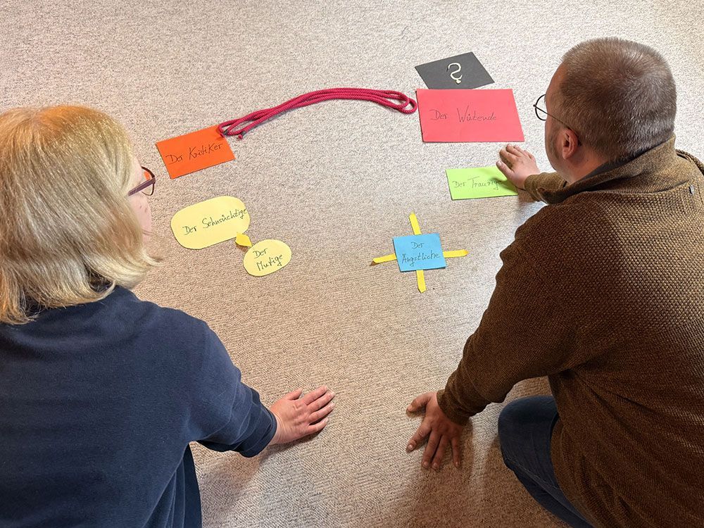 Zwei Personen knien auf dem Boden und arrangieren bunte Haftnotizen, die mit einer roten Schnur verbunden sind.