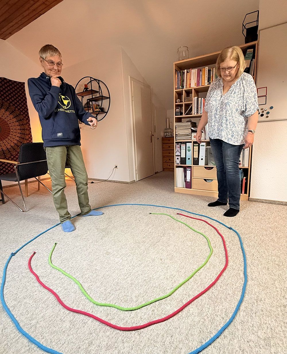 Zwei Personen stehen neben bunten Bodenschlaufen in einem Raum, eine von ihnen hält ein Seil oder eine Schnur.