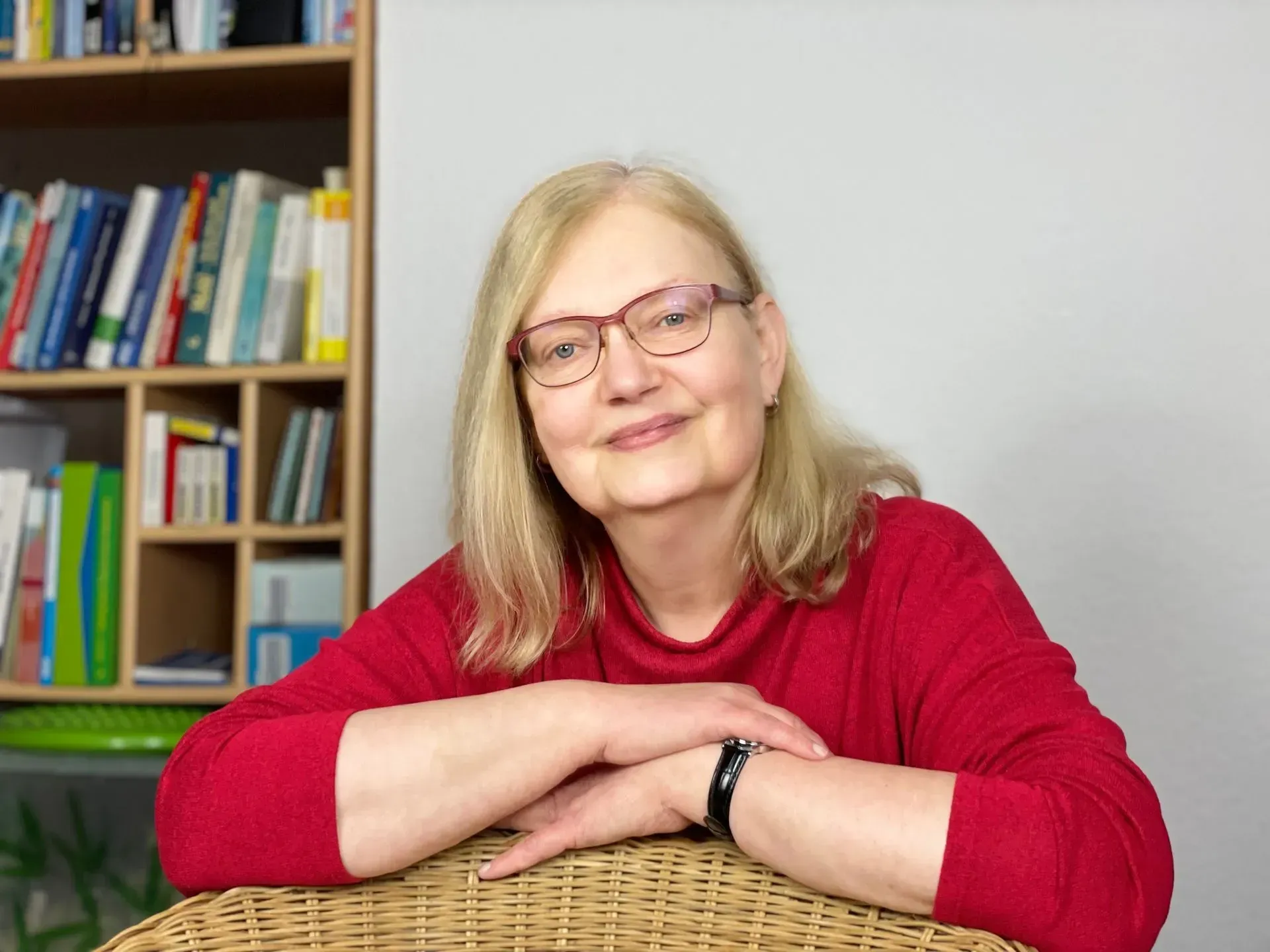 Frau in einem roten Pullover lehnt lächelnd an einem Schreibtisch in einem Zimmer mit Bücherregalen.