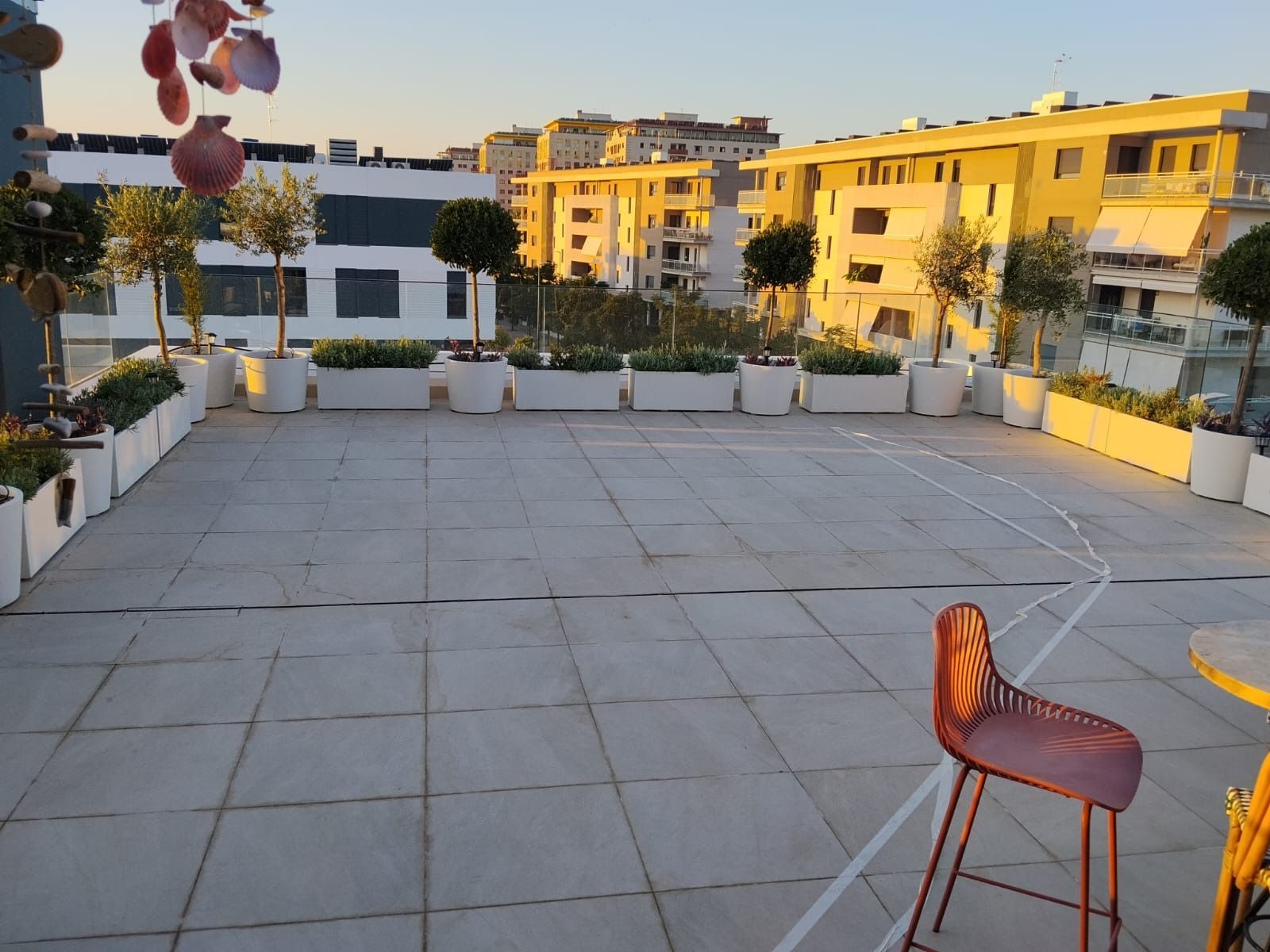 Terraza vacía en la azotea con suelo de baldosas, una silla naranja, plantas en macetas y edificios amarillos al atardecer.