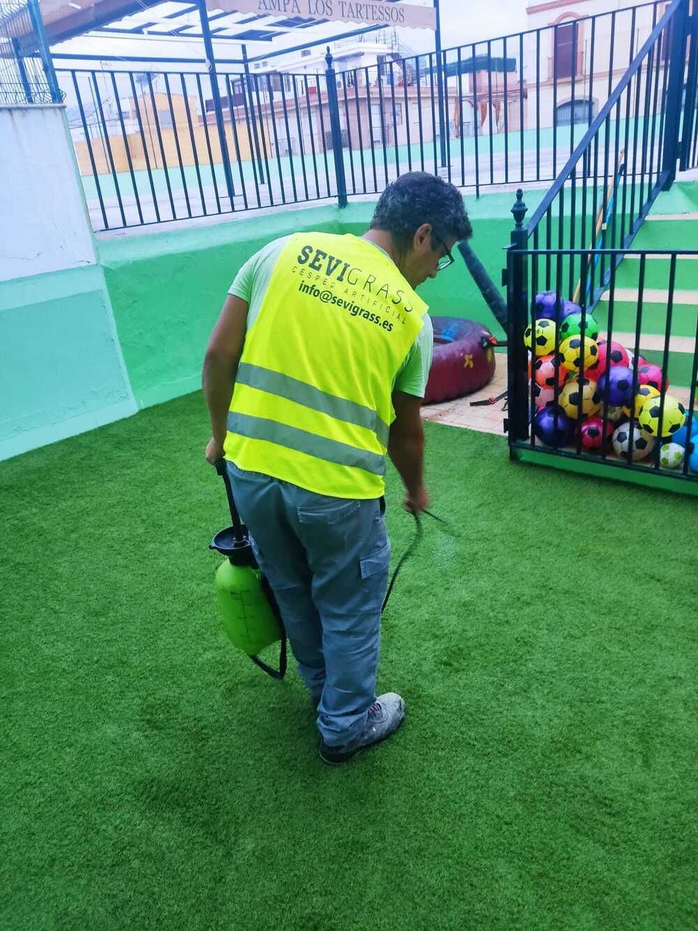 Una persona con un chaleco amarillo se inclina cerca de un contenedor de pelotas de colores sobre césped verde en un área de juegos cercada.