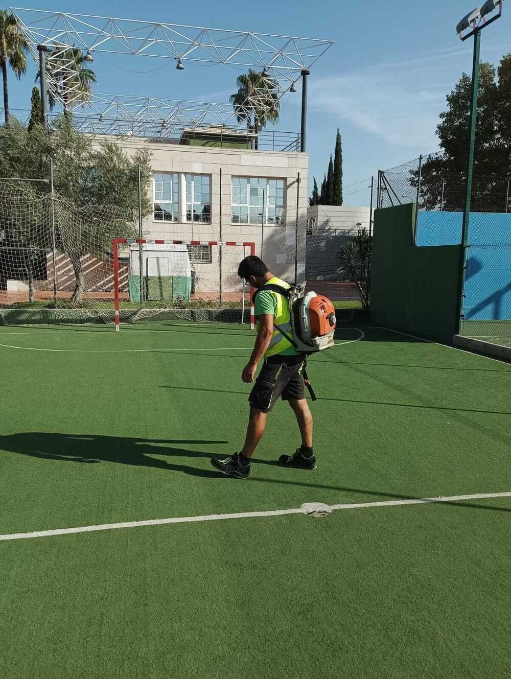 Una persona con un chaleco de neón y una mochila camina por un campo de fútbol al aire libre cerca de una portería.