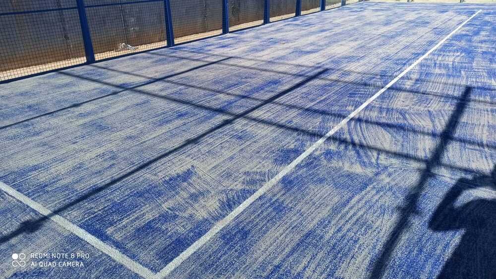 Cancha exterior azul con largas sombras proyectadas sobre una superficie de juego y una valla iluminadas por el sol.