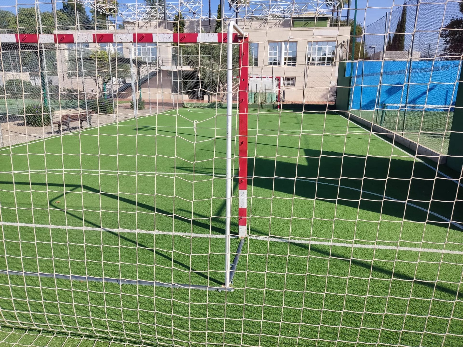 Portería de fútbol con red blanca en un campo de césped artificial verde, con edificios y una lona azul al fondo.
