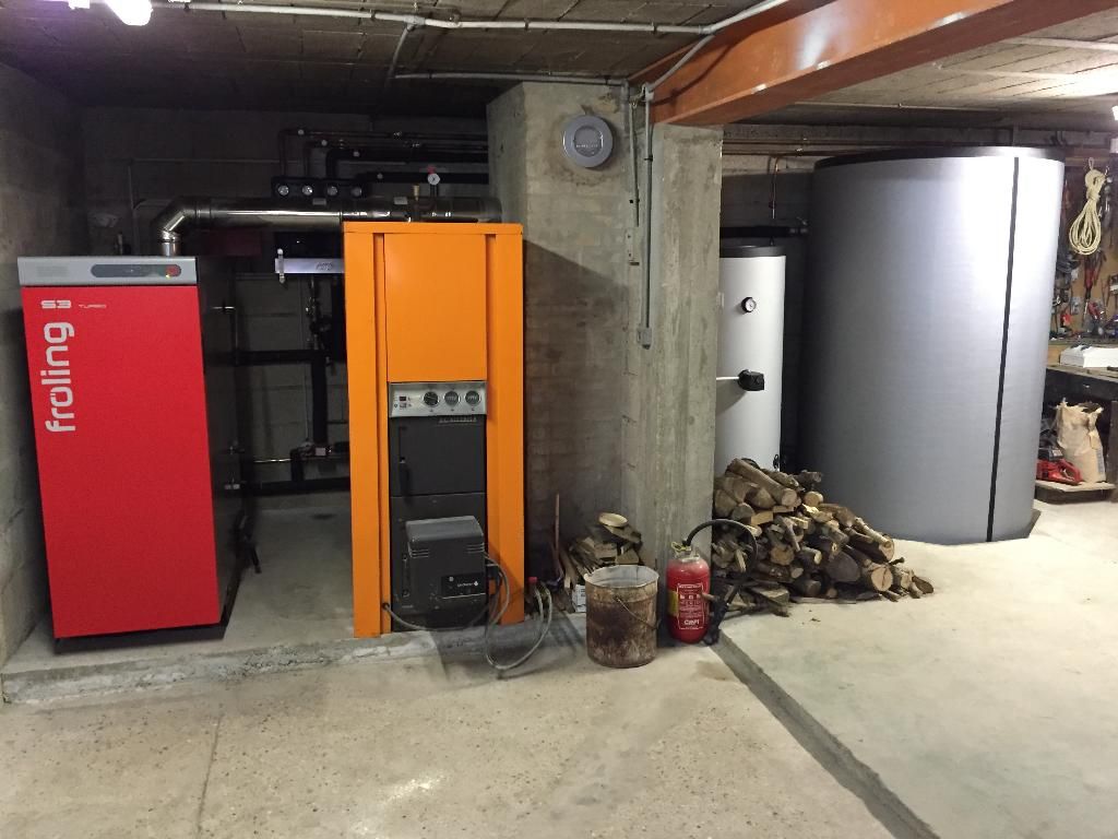 Chaufferie avec unités de chauffage rouges et oranges, un grand réservoir d'eau et du bois de chauffage empilé au sous-sol.