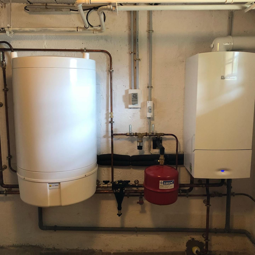 Local technique avec un réservoir blanc, une chaudière, un vase d'expansion rouge et des tuyaux apparents sur un mur.