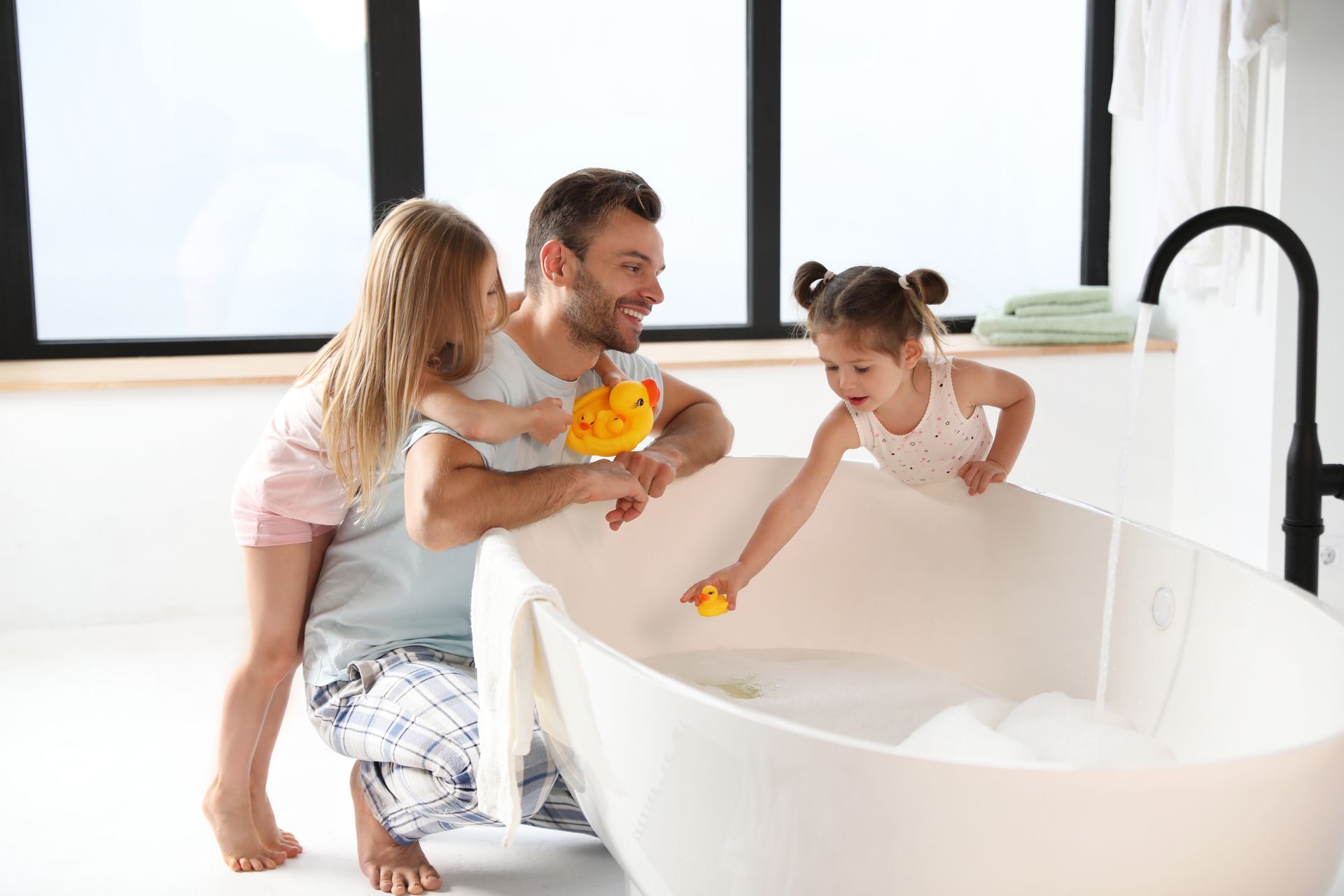 Une famille donne le bain à un enfant dans une baignoire blanche près d'une fenêtre lumineuse, un parent tenant un canard en plastique jaune.