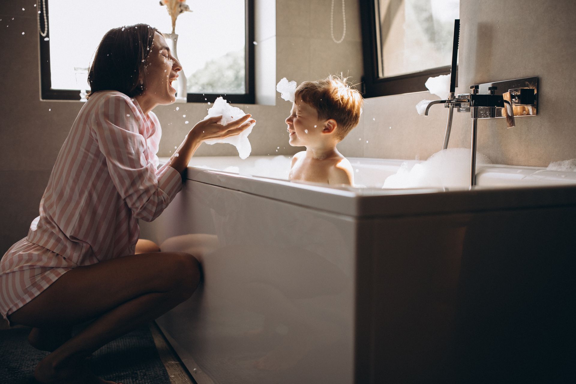 Une femme baigne un bébé dans une baignoire blanche près d'une fenêtre ensoleillée.