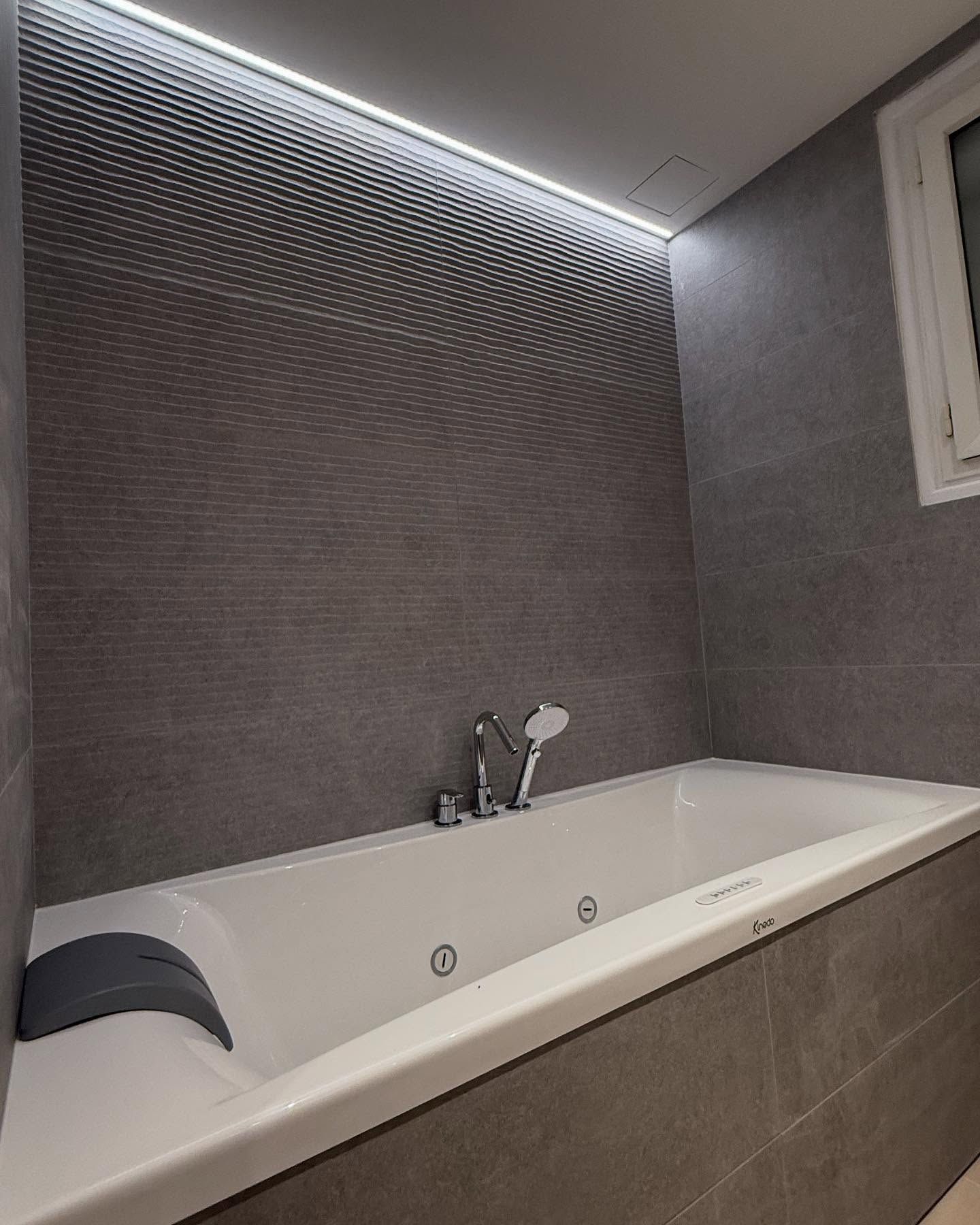 Salle de bains moderne grise avec baignoire blanche, mur en mosaïque et bande LED éclairée au plafond.