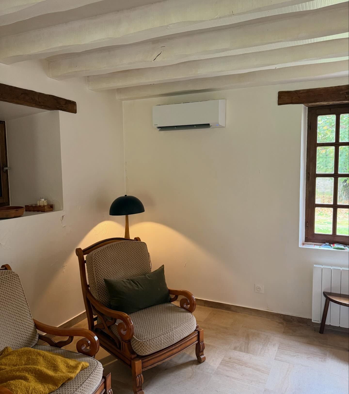 Salon confortable avec fauteuils en osier, lampe verte, murs blancs, poutres apparentes et une fenêtre laissant entrer la lumière du jour.