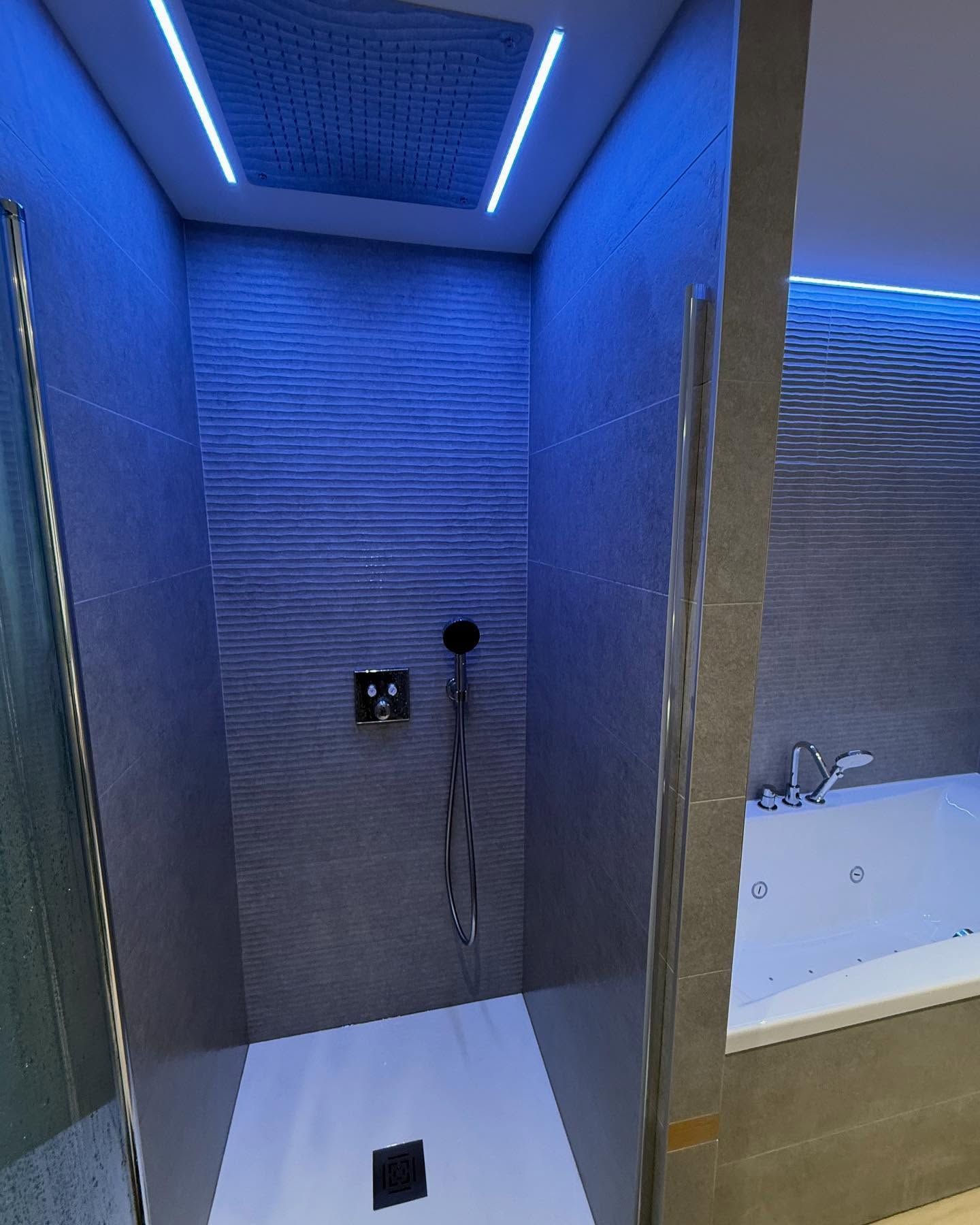 Coin douche moderne éclairé en bleu à côté d'une baignoire avec pommeau de douche mural et murs carrelés.