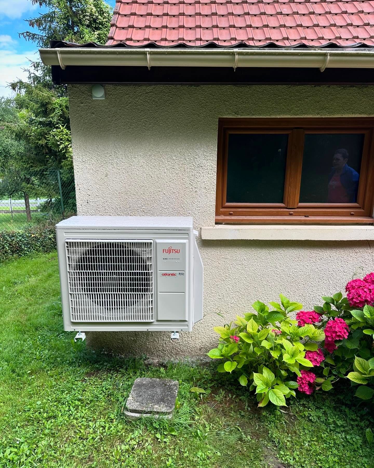 Climatiseur mural sous une fenêtre à l'extérieur d'une maison, à côté d'une pelouse verte et de fleurs roses.
