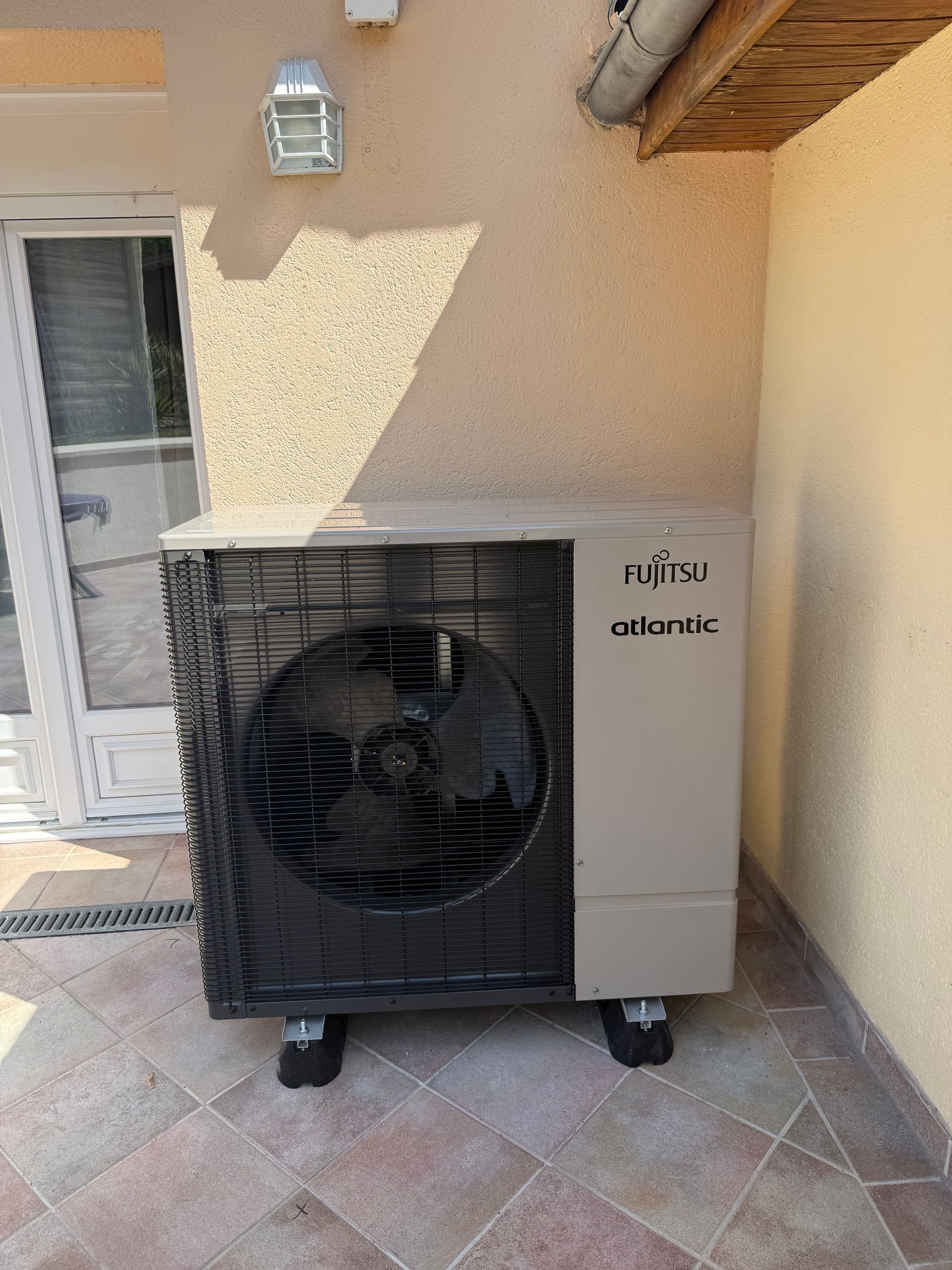 Unité de climatisation extérieure à côté d'un mur et d'une porte en stuc beige sur une terrasse carrelée.