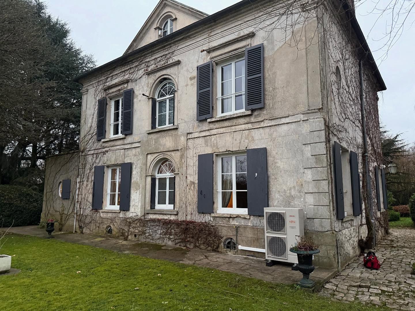 Vieille maison en pierre avec des volets gris, des fenêtres cintrées et une cour gazonnée.