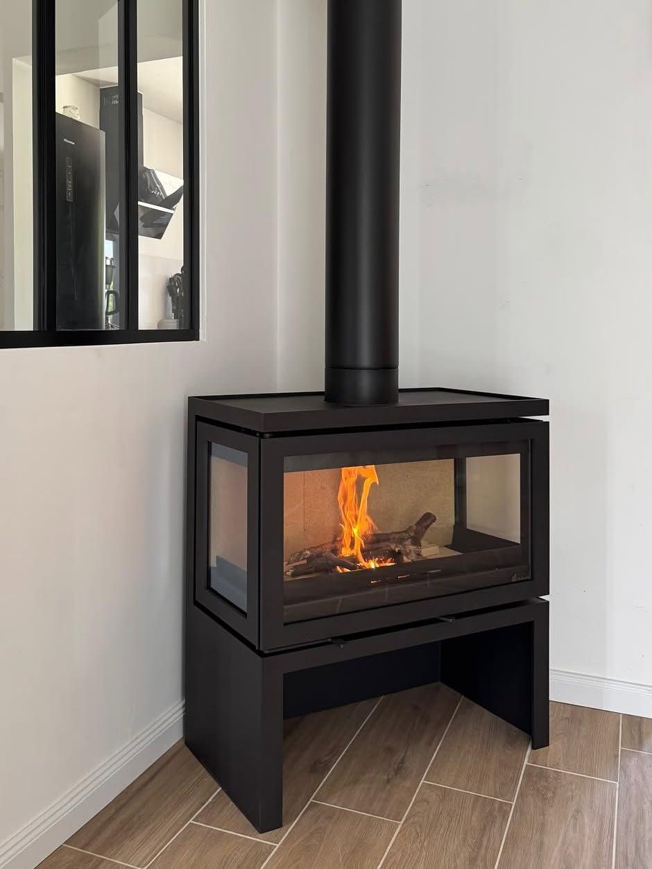 Poêle à bois noir avec foyer vitré, flammes orange allumées, dans une pièce d'angle lumineuse.
