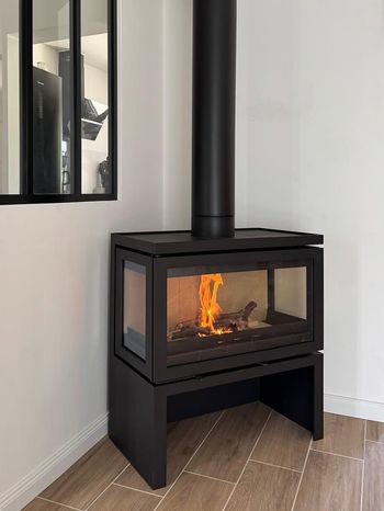 Poêle à bois noir avec foyer vitré, flammes orange allumées, dans une pièce d'angle lumineuse.