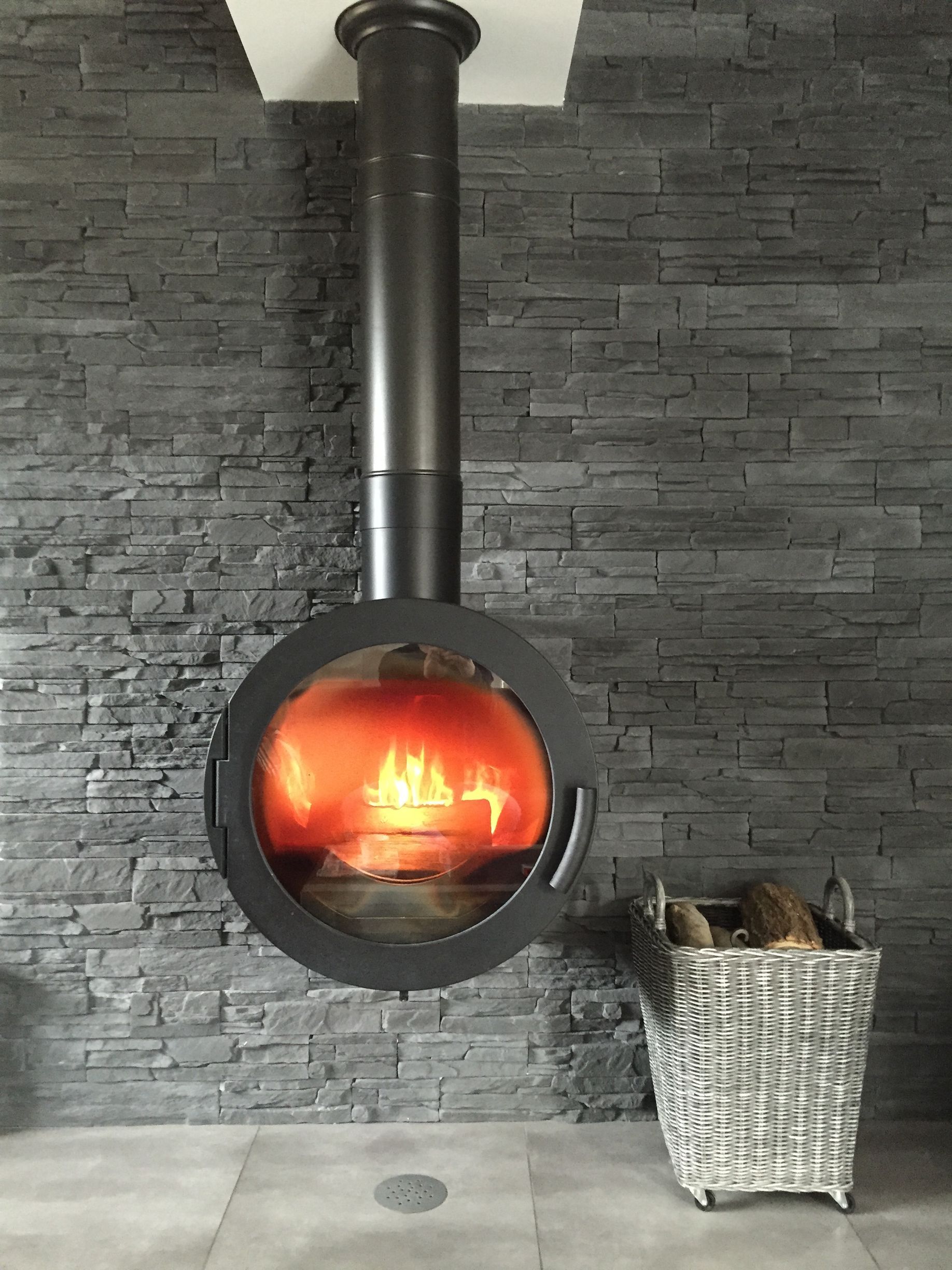 Cheminée ronde moderne avec un feu crépitant contre un mur de pierre sombre, à côté d'un panier en osier rempli de bois de chauffage.