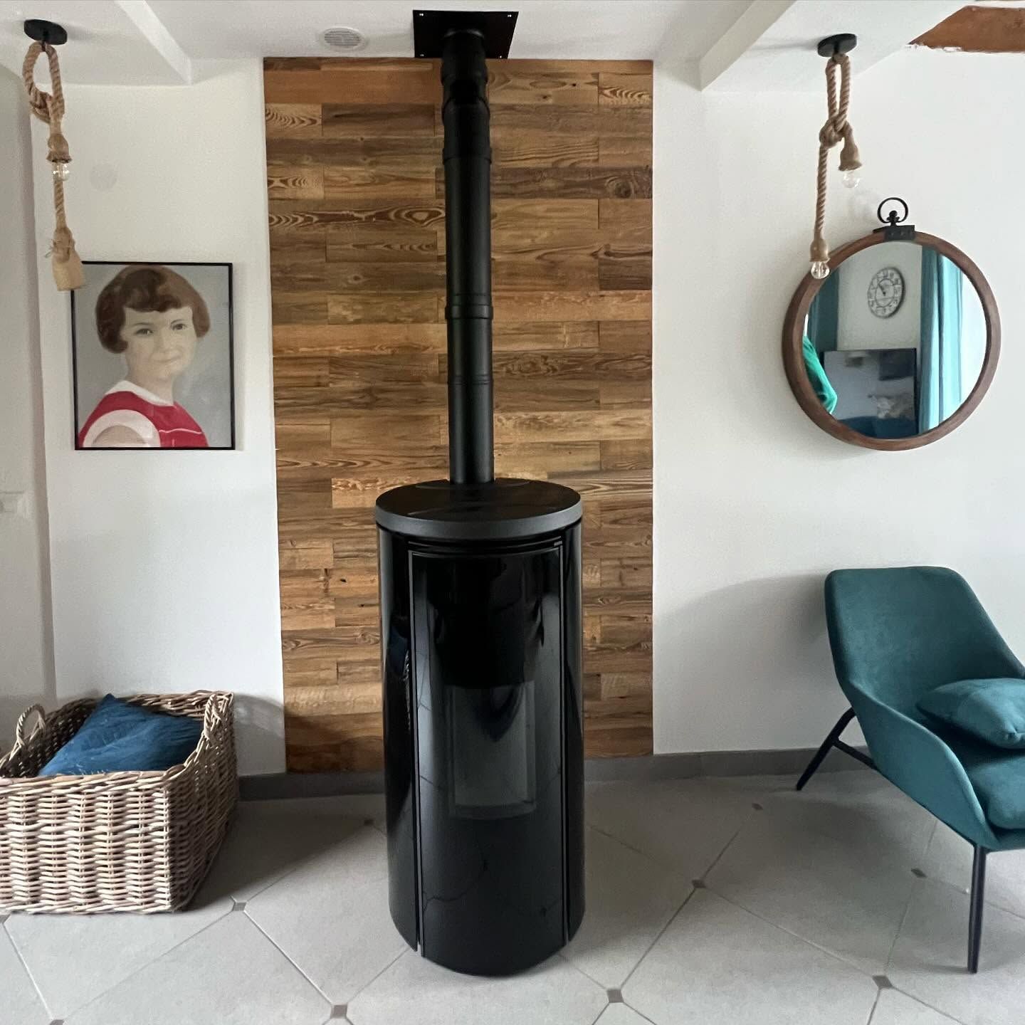 Salon moderne avec un poêle à bois cylindrique noir adossé à un mur en bois, flanqué d'un panier et d'une chaise turquoise.