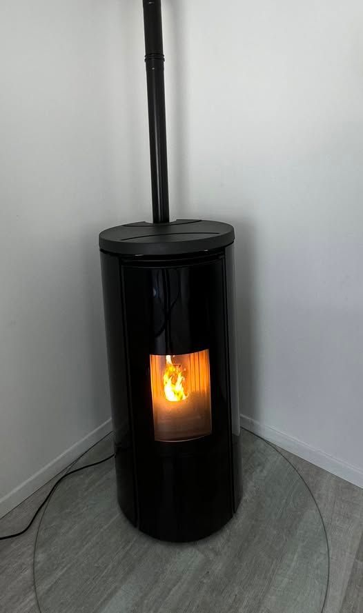 Poêle à granulés cylindrique noir avec un feu incandescent, placé dans un coin sur un sol carrelé.