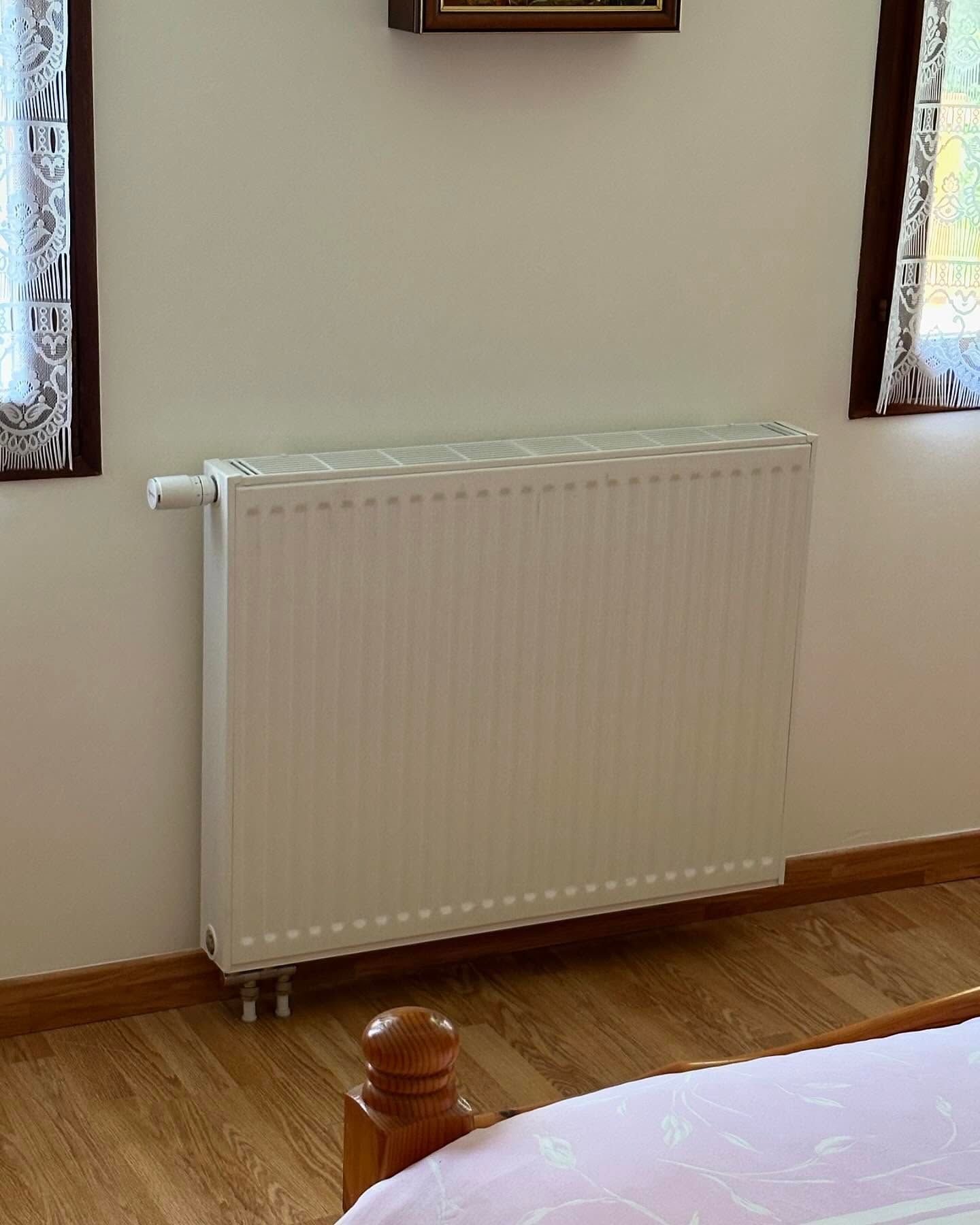 Radiateur blanc fixé sur un mur beige entre deux fenêtres dans une pièce au parquet.