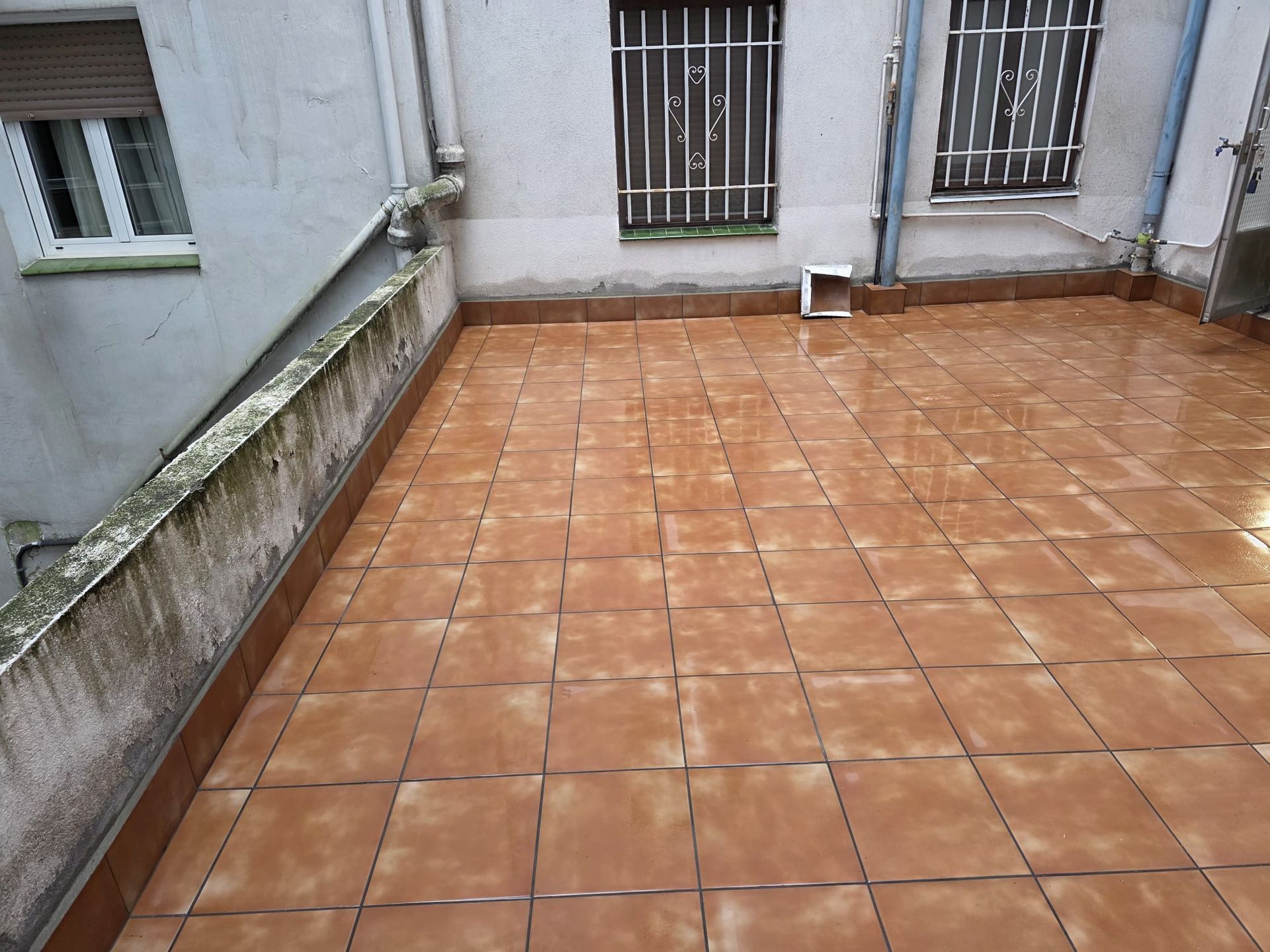 Vista desde un ángulo elevado de un suelo de terraza embaldosado en color terracota, flanqueado por un muro bajo de hormigón desgastado y las paredes de una casa.