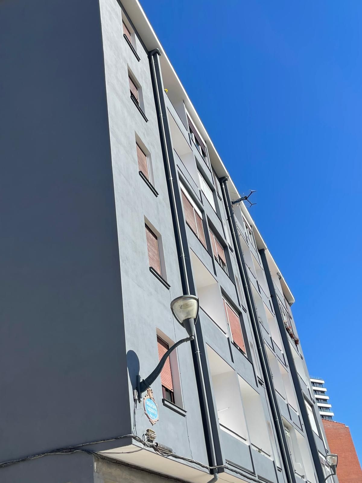 Un edificio de apartamentos alto, de color gris claro, que se recorta contra un cielo azul despejado, con tuberías verticales oscuras y una farola.