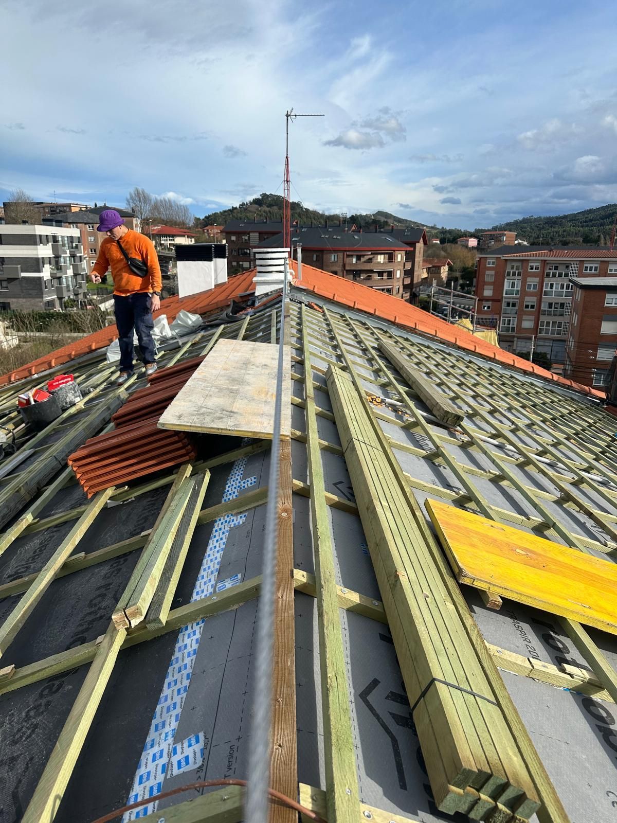 En un día soleado, una persona con una chaqueta naranja trabaja en un tejado en construcción, con estructura de madera y tejas.