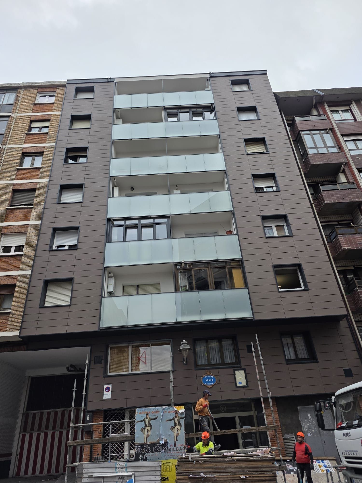 Obreros de la construcción trabajan frente a un edificio de apartamentos de varias plantas con una moderna fachada gris oscuro y balcones de cristal.