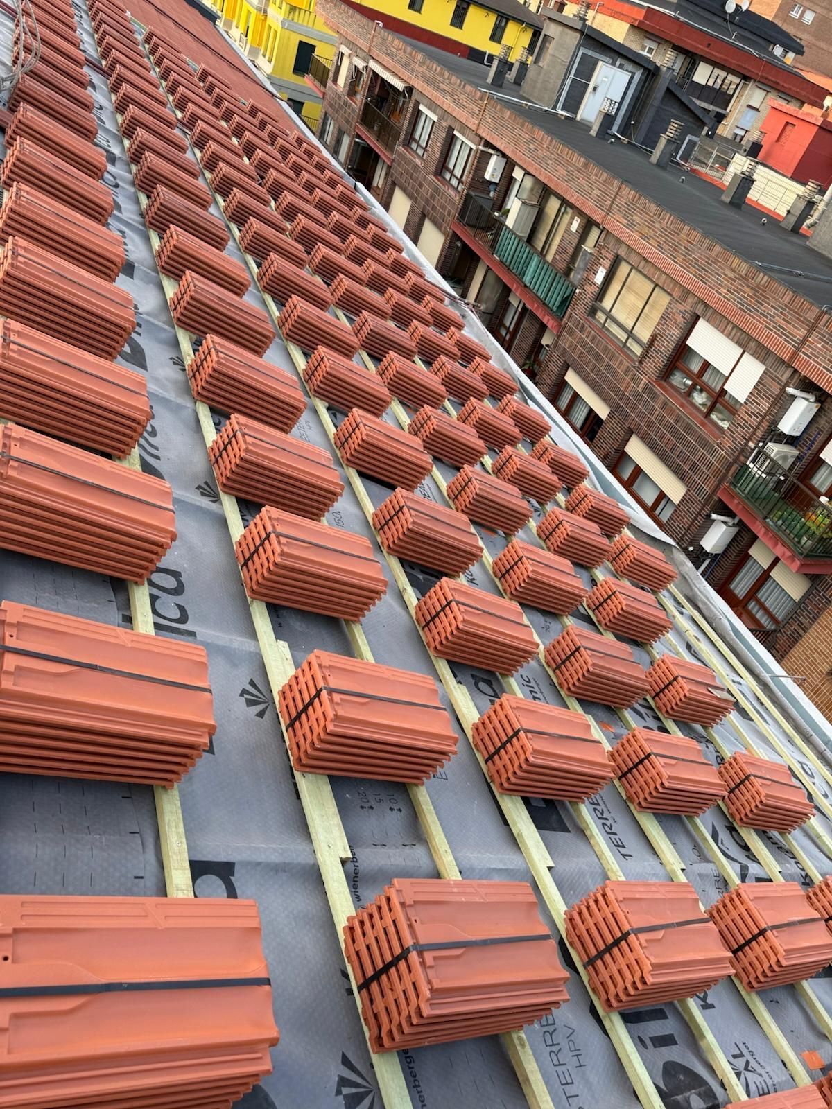 Hileras de tejas de arcilla roja apiladas recubren un tejado inclinado cubierto con una capa protectora y listones de madera.