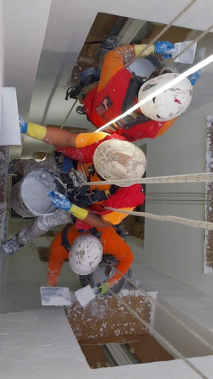 Trabajadores con uniformes y cascos naranjas de alta visibilidad descienden en rápel por la fachada de un edificio, pintando las paredes blancas.