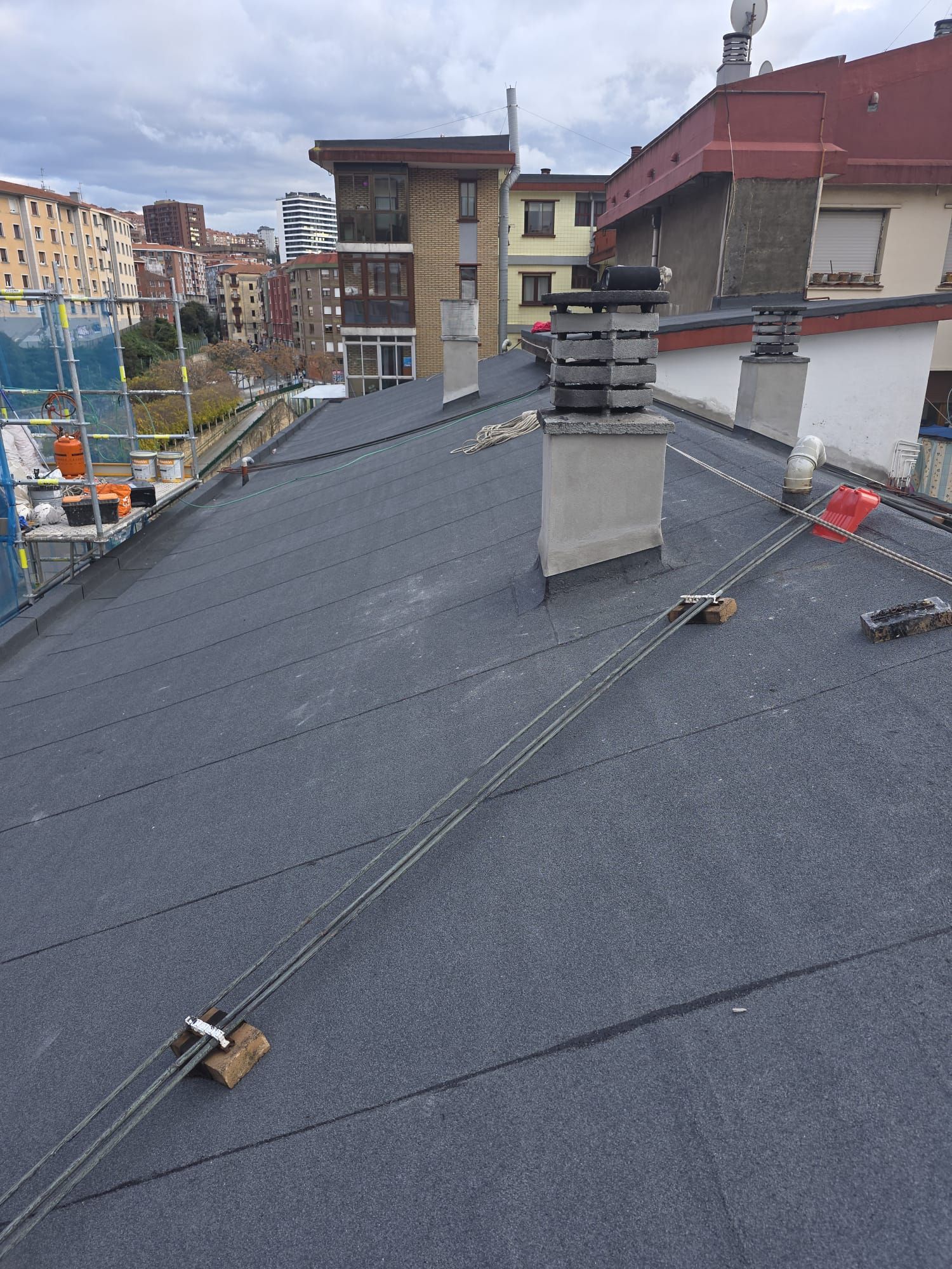 Una azotea cubierta de un material gris y texturizado, con chimeneas, cables y edificios residenciales cercanos bajo un cielo nublado.