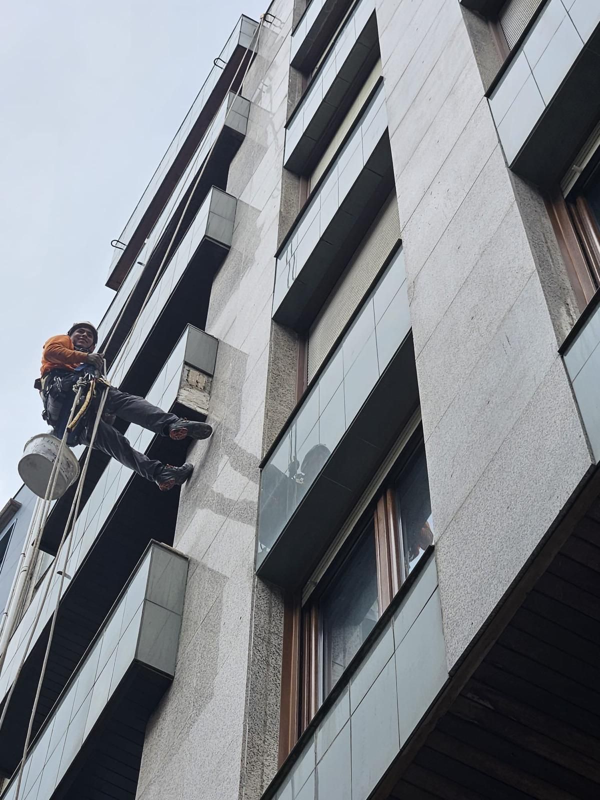 Un obrero de la construcción, suspendido por cuerdas, limpia la fachada de un alto edificio de apartamentos de color gris.