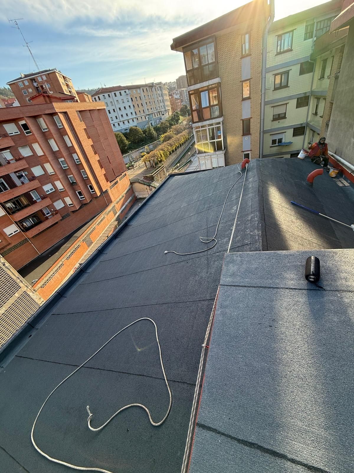 Vista desde un ángulo elevado de un tejado plano en reparación, con membrana bituminosa oscura, cuerda y un trabajador visibles al fondo.