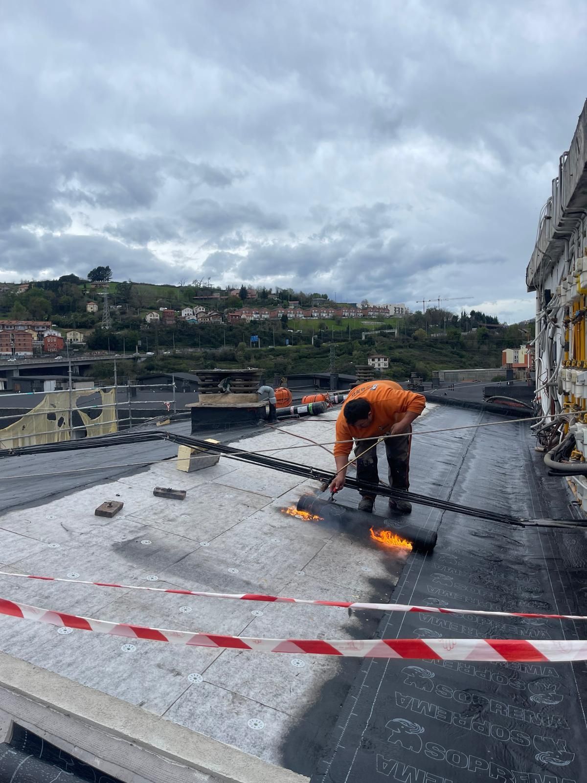 Un trabajador con una camisa naranja utiliza un soplete para instalar material de techado bituminoso impermeable en el techo plano de un edificio.