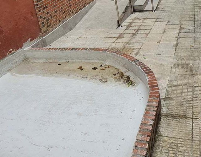 Una plataforma de hormigón con un borde curvo de ladrillo, adyacente a una pasarela pavimentada y una rampa para sillas de ruedas junto a una pared de ladrillo rojo.