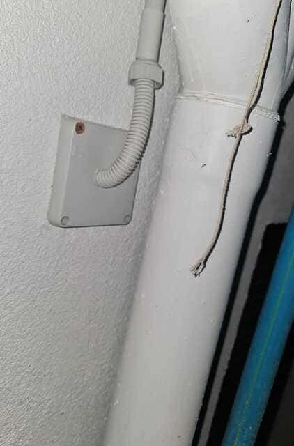 Una pared blanca con una caja de conexiones eléctricas cuadrada de color gris, un conducto flexible blanco y una tubería grande pintada de blanco.