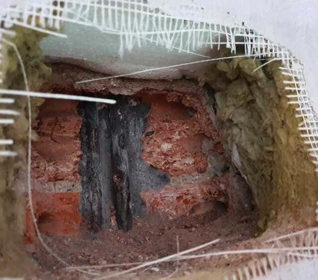Un agujero en una pared exterior deja al descubierto ladrillos rojos, un relleno o mortero oscuro vertical y un aislamiento circundante con malla metálica.