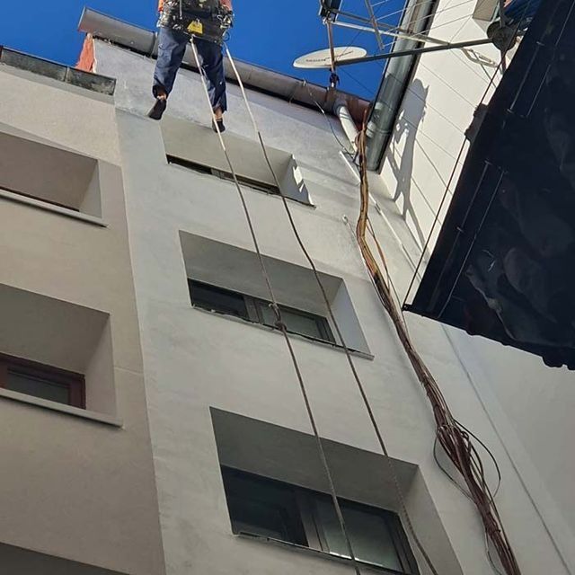 Una persona desciende en rápel por el lateral de un edificio blanco alto, cerca de una antena parabólica y un gran manojo de cables.