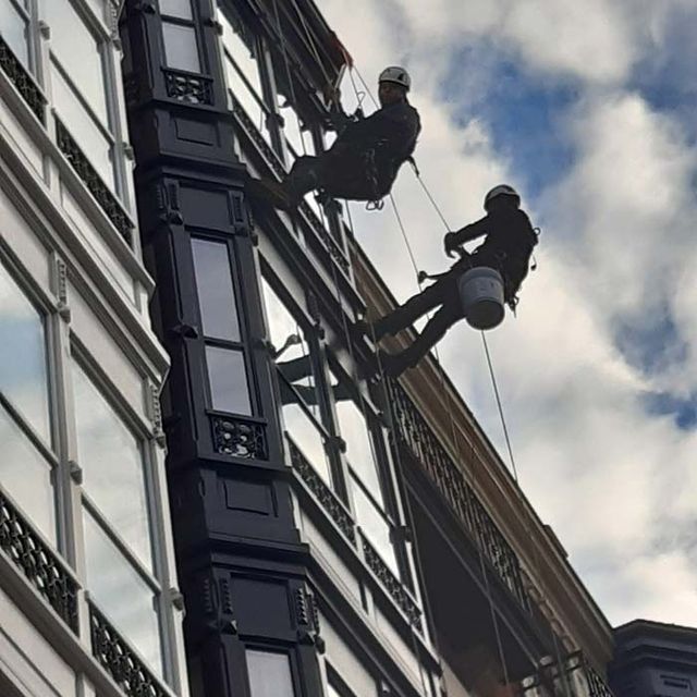 Dos trabajadores con cascos y arneses descienden en rápel por la fachada de un edificio para limpiar sus ventanas.