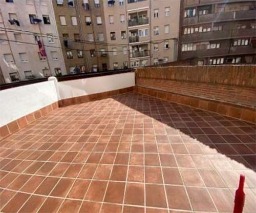 Una terraza en la azotea con suelo de baldosas marrones, rodeada de paredes blancas y de ladrillo, con vistas a un complejo de apartamentos urbanos.