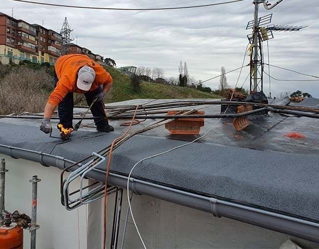 Una persona con una chaqueta naranja instala material de techado en un tejado plano, con una torre de servicios públicos y cables al fondo.