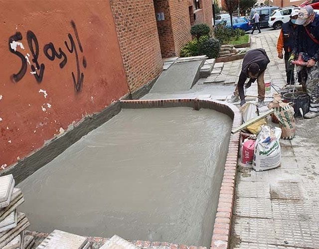 Los trabajadores vierten hormigón para crear una rampa y una plataforma accesibles frente a un edificio con grafitis en la pared.