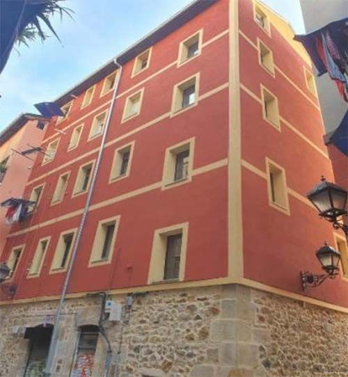 Un edificio de varias plantas con fachada de color rojo óxido, detalles en color crema y base de piedra, visto desde un ángulo bajo a nivel de la calle.