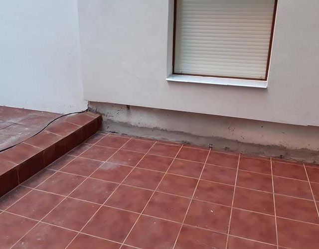 Un patio con baldosas rojas y un escalón elevado, que da paso a una pared blanca con una ventana y una pequeña junta de mortero sin terminar.