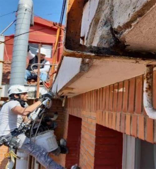 Dos trabajadores con equipo de seguridad reparan una cornisa de hormigón dañada en el exterior de un edificio de ladrillo.