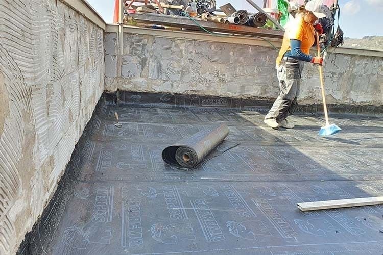 Un trabajador utiliza una escoba para barrer una azotea plana cubierta con rollos de membrana impermeable negra.