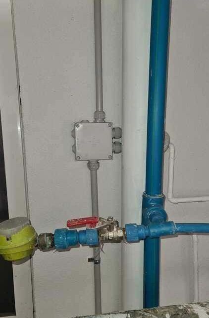 Un contador de agua y una válvula de cierre conectados a tuberías azules en una pared blanca con una caja de conexiones eléctricas encima.