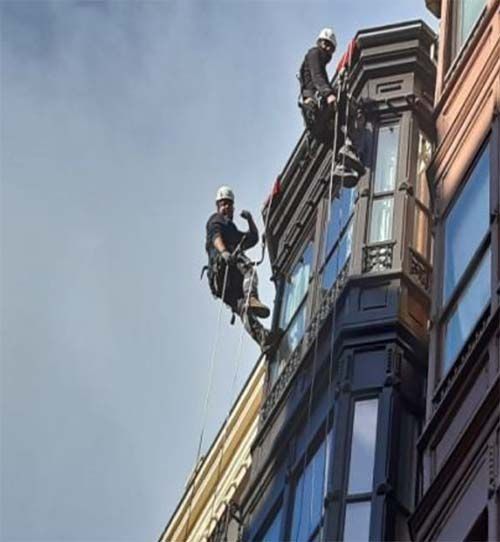 Dos limpiadores de ventanas, equipados con cascos y arneses de seguridad, descienden por la fachada de un edificio alto utilizando cuerdas.