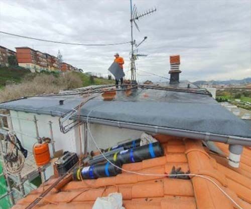 Un trabajador con ropa de color naranja brillante instala material de cubierta gris impermeable en un tejado plano junto a un tejado de tejas.